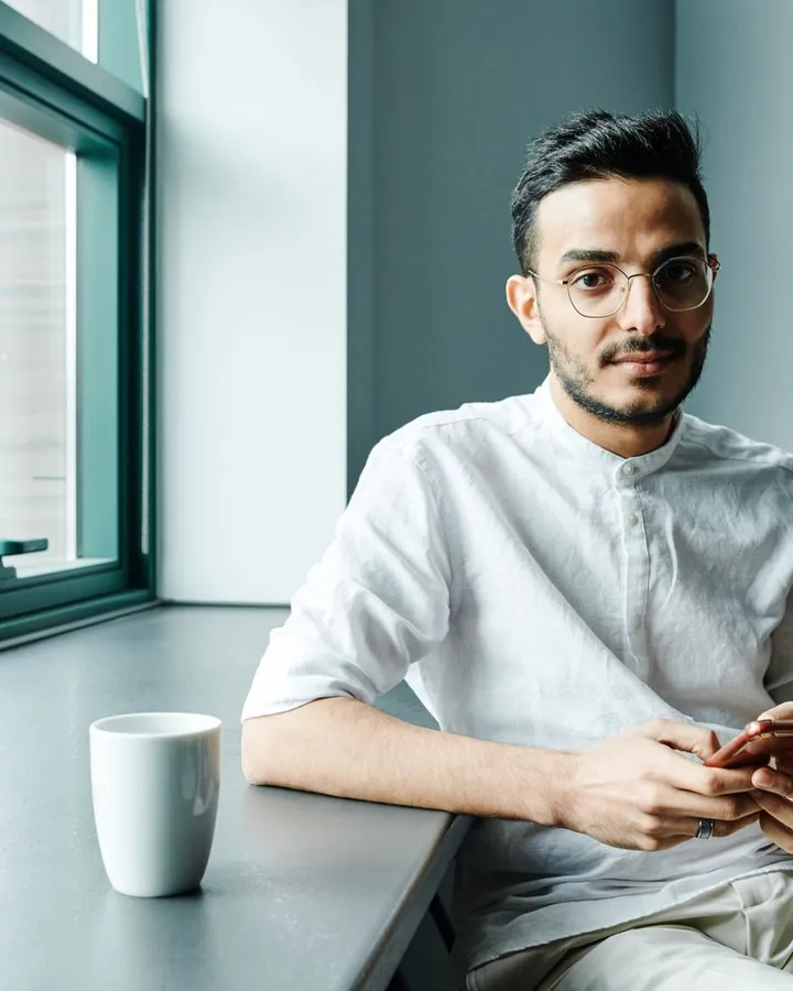 Mann mit Brille und weißem Leinenhemd sitzt allein am Fenstertresen eines hellen Cafés, Handy in der Hand, Kaffeebecher neben sich