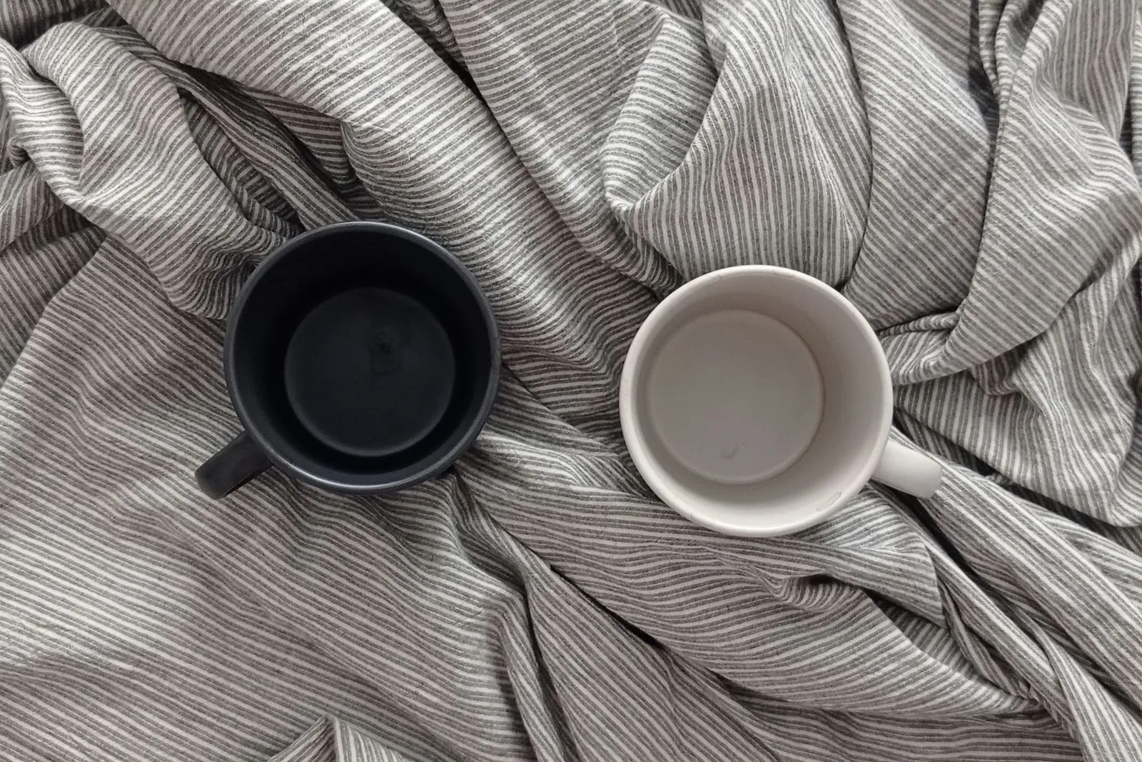 Zwei leere Kaffeebecher, einer schwarz, einer weiß, auf zerwühlter grau gestreifter Bettwäsche, von oben fotografiert