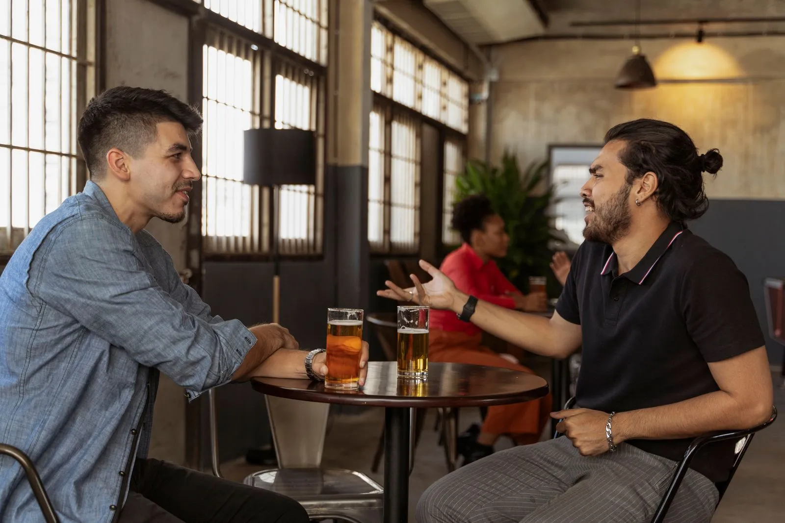 Zwei Männer sitzen sich in einer hellen Bar mit hohen Sprossenfenstern an einem kleinen runden Tisch gegenüber, vor ihnen zwei Gläser Bier, einer erzählt mit ausgestreckter Hand, der andere hört lächelnd zu