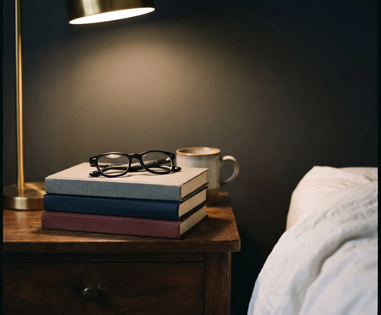 Drei Bücher mit Lesebrille auf einem Nachttisch aus dunklem Holz, daneben eine Messinglampe, eine Tasse und die Bettkante, warmes Abendlicht
