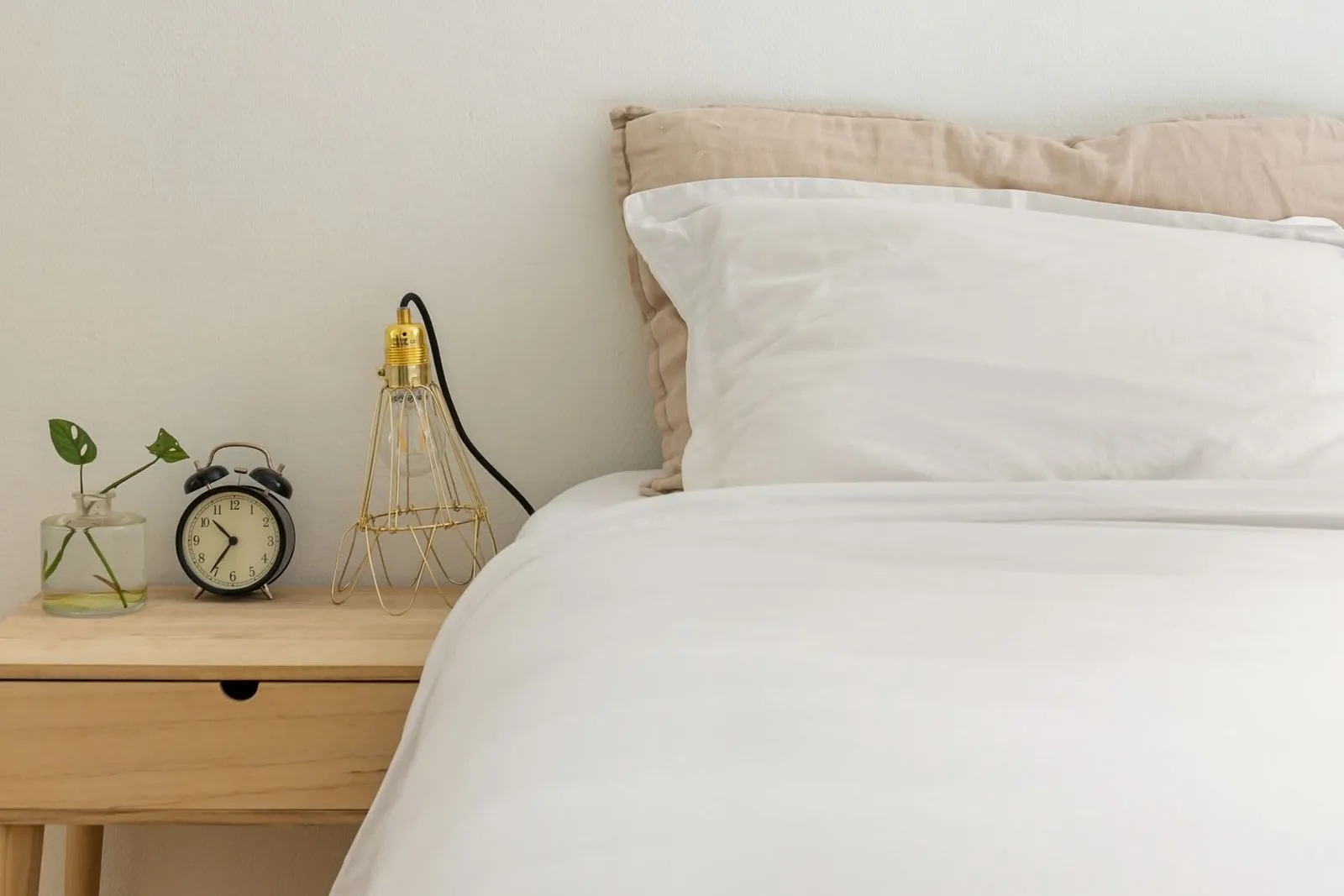 Aufgeräumtes Schlafzimmer bei Tageslicht, weiße Bettwäsche vor einem beigen Leinenkissen, daneben ein Holznachttisch mit Drahtlampe, Wecker und einem Steckling im Glas