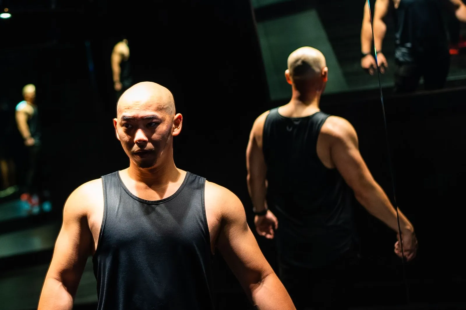 Muskulöser Mann im schwarzen Tanktop in einem dunklen Trainingsraum, hinter ihm wirft eine Spiegelwand sein Bild mehrfach zurück