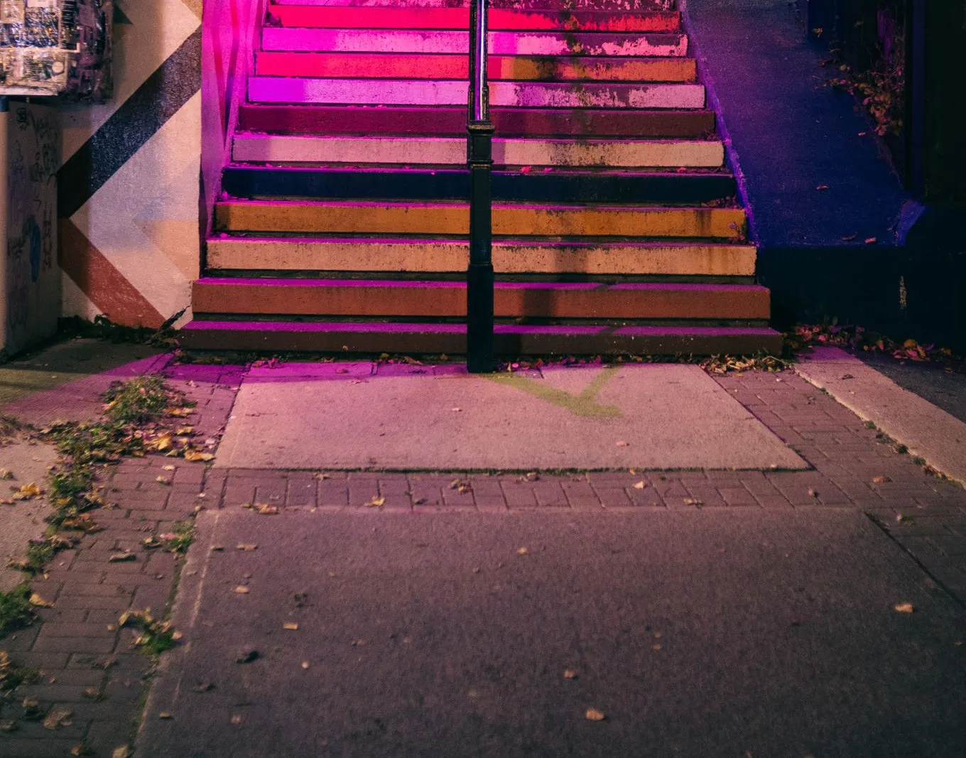Leere Treppe mit abgewetzten Regenbogenstufen bei Nacht, magentafarbenes Licht, Herbstlaub auf dem Gehweg